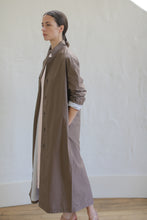 Load image into Gallery viewer, Dressed in a long Clay-colored Painter's Coat with buttons and pockets, a person stands sideways against a light-colored wall. Their hair is tied back, and the background features a wooden floor and a white, curved baseboard.