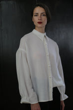 Load image into Gallery viewer, A person with short, dark hair stands against a dark background, wearing a white, long-sleeved button-up Poet Shirt - Japanese Wool Gauze | Undyed with a slight sheen and loose fit. The expression is neutral, and they have red lipstick. The overall mood of the image is minimalist and elegant, showcasing sustainable design.