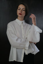 Load image into Gallery viewer, A person wearing a loose, long-sleeved Poet Shirt - Japanese Wool Gauze | Undyed with pearl buttons stands against a dark background. They have short dark hair and are posing with one hand raised near their face, gazing into the distance with a neutral expression.