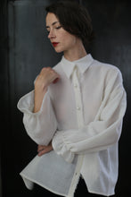 Load image into Gallery viewer, A person with dark hair is standing against a dark background, wearing a loose-fitting, long-sleeved Poet Shirt - Japanese Wool Gauze | Undyed with buttons. They have a thoughtful expression while looking down and resting one hand on their neck. Their red lipstick adds a pop of color.