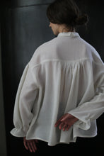 Load image into Gallery viewer, A person with dark hair styled in a bun is seen from the back, wearing a loose-fitting, white Poet Shirt - Japanese Wool Gauze | Undyed. The person has one hand behind their back while standing against a dark, blurred background. The blouse appears to be crafted from Japanese wool gauze, emphasizing sustainable design.