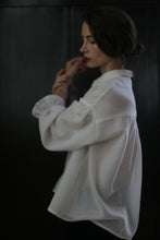 Load image into Gallery viewer, A woman with dark hair is standing sideways against a dark background. She is wearing an oversized Poet Shirt - Japanese Wool Gauze | Undyed, its long sleeves draping elegantly. Her hair is loosely tied back, and she appears to be lost in thought, with her hands gently touching her face.