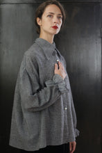 Load image into Gallery viewer, A woman with brown hair stands in front of a dark background, wearing the Poet Shirt - Japanese Wool Gauze | BW Houndstooth. She gazes slightly upward with a serious expression, resting her left hand on her chest, exuding a Byronesque flair.