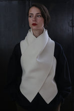 Load image into Gallery viewer, A person poses against a dark background, wearing a black top and a cream-colored, scarf-like garment draped around the neck and chest in a cross-over fashion. The X Scarf - Double-face Cashmere | Undyed features ceramic buttons as accents. They have short, straight hair and wear red lipstick, looking slightly off to the side.
