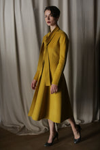 Load image into Gallery viewer, Against luxurious, flowing curtains, a person exudes elegance in a Saffron Doubleface Cashmere Ballerina Coat with a matching skirt and high heels. Soft lighting casts shadows on the textured fabric, highlighting the outfit's shape and color.