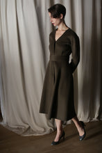 Load image into Gallery viewer, A person wears The Ballerina Coat in Doubleface Cashmere Olive with its elegant silhouette, standing on a wooden floor in black heels. They have short, dark hair, set against a backdrop of draped light-colored fabric.