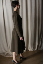 Load image into Gallery viewer, Against light-colored drapes, a person stands in profile, wearing The Ballerina Coat. This luxury coat, featuring a tailored silhouette and crafted from olive double-faced cashmere, pairs elegantly with dark shoes as gentle lighting casts soft shadows.