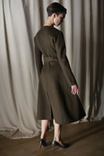 Load image into Gallery viewer, Wearing "The Ballerina Coat | Doubleface Cashmere Olive," a person showcases its luxurious, tailored silhouette with long sleeves and a back tie. Their short hair and high heels complement the look, set against a curtained background.