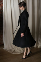 Load image into Gallery viewer, A person wearing a high-heeled luxury fashion ensemble with The Ballerina Coat in Doubleface Cashmere Black is captured in profile, standing before a light-colored curtain alongside a partially visible brick wall.