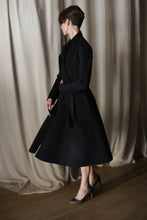 Load image into Gallery viewer, In a dimly lit room with soft, draped curtains, a person elegantly stands sideways with short hair, showcasing The Ballerina Coat in black Doubleface Cashmere and gray high-heeled shoes, epitomizing luxury fashion with its tailored silhouette and flowing design.