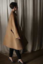 Load image into Gallery viewer, Wearing the Kyoto | Camel luxury women’s double-faced cashmere coat with black pants, a person walks indoors by cream-colored curtains. Soft lighting highlights the coat as she is seen in profile.