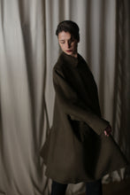 Load image into Gallery viewer, In dim light, a person with dark hair and a Kyoto | Olive coat stands lightly turned before a cream curtain, casting soft shadows. The lighting accentuates the voluminous coat's shape, creating a dramatic silhouette and moody ambiance.