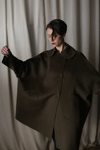 Load image into Gallery viewer, A person stands with a neutral expression against a draped curtain backdrop, the oversized Kyoto Olive coat creating a dramatic silhouette as they hold their arms outstretched.