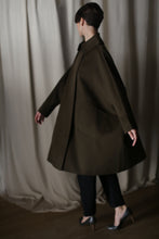 Load image into Gallery viewer, A person with short hair models the Kyoto | Olive oversized coat, featuring a dramatic silhouette and large pockets, as they walk leftward. They pair it with black pants and metallic heels, set against a backdrop of beige curtains that create a soft, elegant atmosphere.