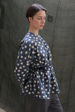 Load image into Gallery viewer, A person with short dark hair stands sideways, looking down, wearing the Qipao Blouse | Polkadot Seersucker Indigo belted at the waist with gray trousers. The background features textured dark green fabric.