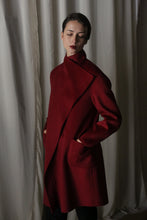 Load image into Gallery viewer, A woman in the Winston Long Double-face coat in wine, with draped collar and pockets, stands against a backdrop of softly draped fabric. Her short dark hair is styled neatly as she gazes downward serenely.