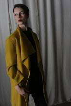 Load image into Gallery viewer, A dark-haired individual wears a Saffron-colored Winston Long Double-Face coat over a black outfit. They gaze left with a neutral expression, as white curtains softly drape in the background, enhancing the dim, moody atmosphere.