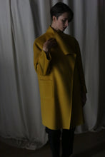 Load image into Gallery viewer, In a dimly lit room with curtain backdrops, a person stands enveloped in a dramatic atmosphere wearing the Winston Long Double-face coat in Saffron. The cashmere coat features a draped collar and two visible pockets, highlighted by soft shadows cast by the lighting.