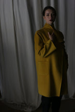 Load image into Gallery viewer, In a dimly lit room, someone leans against soft, white fabric wearing the Winston Long Double-face coat in Saffron. The draped collar and gentle shadows add an air of elegance and mystery to the moody ambiance.