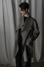 Load image into Gallery viewer, Wearing the Winston Long Double-face coat in undyed natural, a person with short hair looks down, hands in pockets over dark clothing—a true wardrobe foundation against soft, light-colored fabric curtains.