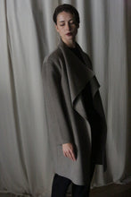 Load image into Gallery viewer, A person stands before a curtain in the Winston Long Double-face | Undyed Natural, a perfect wardrobe foundation. Dim lighting casts shadows as they gaze slightly downward with short hair, embodying timeless neutrals.