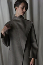 Load image into Gallery viewer, Modeling short hair and the Winston Long Double-face coat in Undyed Natural, an enduring neutral piece suitable for any wardrobe, they gently touch the high collar against a softly lit backdrop, exuding a pensive elegance.