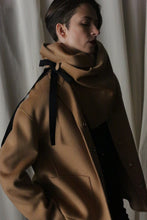 Load image into Gallery viewer, A person wearing a tan coat with cashmere-covered buttons and an X Scarf - Double-face Cashmere in Camel stands against a light curtain backdrop. They have short, dark hair, one hand in their pocket, looking slightly downward in dramatic, moody lighting.