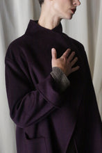 Load image into Gallery viewer, Against a light curtain, a person wears the Winston Long Double-face coat in aubergine with a high collar made of double-faced cashmere. Their hand rests on their chest, and they have short hair. Soft lighting enhances the calm atmosphere, highlighting the coat's versatile styling options.