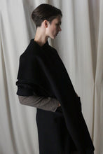 Load image into Gallery viewer, A person with short hair stands in profile against a cream-colored curtain, draped in the Winston Long Double-face | Black shawl made of double-faced cashmere over a light long-sleeve top. The soft lighting creates shadows for a minimalist and contemplative feel.