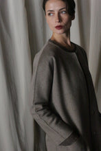 Load image into Gallery viewer, A person with short dark hair stands contemplatively, wearing the Battersea Double-Face Cashmere in Undyed Natural. The coat's versatile silhouette and side pockets create a minimalist aesthetic complemented by light-colored curtains in the background.