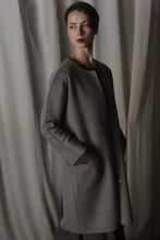 Load image into Gallery viewer, Against a softly draped fabric backdrop, a person wears the Battersea Double-Face Cashmere coat in undyed natural, exuding luxury. With hands in pockets and a contemplative look, they stand in soft lighting that enhances the coat's versatile silhouette and creates an elegant atmosphere.