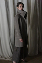 Load image into Gallery viewer, In dim lighting with flowing curtains in the background, a person elegantly poses in profile wearing the Stamford Long coat in Undyed Natural and a dark skirt, gazing towards the camera.