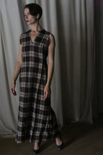 Load image into Gallery viewer, A person with short dark hair stands before draped curtains, wearing The Winter Dress in Japanese Wool Gauze Taupe Plaid with black shoes. The sleeveless, long dress features brown and black plaid shades. They look to the side neutrally.