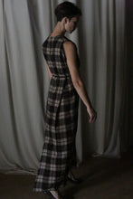 Load image into Gallery viewer, A person with short hair stands sideways in The Winter Dress | Japanese Wool Gauze Taupe Plaid and black heels, posing against draped, light curtains in dim lighting.