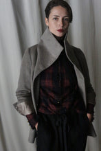 Load image into Gallery viewer, A person stands confidently against a gray curtain, wearing the Stamford II in Undyed Natural over a red plaid shirt and dark pants. They have short hair, gaze into the camera with a slight smile, and keep their hands in their pockets, enhancing their poised demeanor.