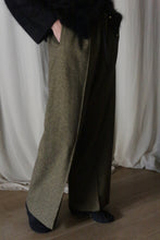 Load image into Gallery viewer, Wearing brushed cotton trousers in moss herringbone from Atelier Lan Jaenicke and a dark top, the person stands gracefully before white draped curtains on a wooden floor, with hands casually resting in pockets, complemented by dark socks.