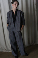 Load image into Gallery viewer, A person stands against a dimly lit curtain in an Atelier Lan Jaenicke suit, featuring a loose-fitting blazer and Brushed Cotton Trousers in blue herringbone. Their hands are in pockets, gazing downward with neatly styled short hair, creating a soft, moody ambiance.