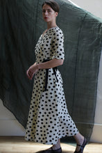 Load image into Gallery viewer, A woman wears the Fitted Wrap Dress | Polkadot Seersucker Ivory, featuring a black-on-ivory polkadot pattern. Indoors on a wooden floor with a dark fabric backdrop, she looks over her shoulder, her hair simply pulled back.