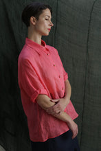 Load image into Gallery viewer, A woman with short brown hair stands in profile, arms folded and eyes closed, wearing the Work Shirt | Ramie Sakura—a loose, coral pink classic ramie shirt with brass buttons—against a dark green fabric backdrop.
