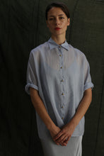 Load image into Gallery viewer, A woman stands before a dark green fabric backdrop, hands folded and expression neutral, wearing the Work Shirt | Ramie Mist—a sheer, light blue ramie button-up.