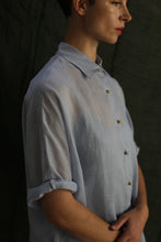 Load image into Gallery viewer, A person in a light blue Work Shirt | Ramie Mist with rolled sleeves and brass buttons stands against a dark background. The photo is cropped above the nose, leaving the person's eyes out of view.