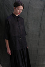 Load image into Gallery viewer, A person with short hair wears the Work Shirt | Ramie Black, featuring solid brass buttons, and a black skirt, standing against a textured dark fabric backdrop and looking downward with a calm expression.