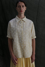 Load image into Gallery viewer, A woman stands against a dark textured background, wearing the Work Shirt | Polkadot Seersucker Butter—a loose, short-sleeved Japanese cotton button-up with brass buttons—paired with a light yellow skirt. She looks at the camera with a neutral expression.