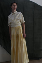 Load image into Gallery viewer, A person stands against a dark fabric backdrop, wearing the Work Shirt in Polkadot Seersucker Butter with rolled sleeves and a long, flowing yellow satin skirt. Soft, subdued lighting creates a moody atmosphere.
