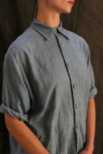 Load image into Gallery viewer, A person in the Work Shirt | Triple Gauze Ai Dye—an indigo-dyed, loose-fit button-up with hand-sewn buttons and rolled sleeves—stands against a dark, textured background, their face partly out of frame.