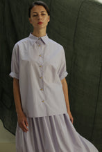 Load image into Gallery viewer, A person stands before a dark green fabric backdrop wearing the Work Shirt | Typewriter Cotton Amethyst, a loose, light lavender Japanese cotton button-up with rolled sleeves, paired with a matching pleated skirt.