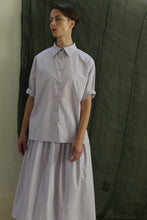 Load image into Gallery viewer, A person stands before a dark green fabric backdrop, wearing the Work Shirt | Typewriter Cotton Amethyst with rolled sleeves and brass buttons, paired with a matching flowy skirt. They gaze to the side, short hair styled neatly.