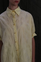Load image into Gallery viewer, A person wears the Work Shirt | Textured Lawn Citrine—a pale yellow, short-sleeved cotton lawn shirt with subtle polka dots and brass buttons—standing against a dark background, their face partially out of frame.