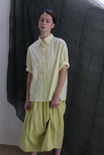 Load image into Gallery viewer, A person with short hair wears the Work Shirt in Textured Lawn Citrine, styled loosely and paired with matching wide-leg pants, standing before a dark, textured fabric backdrop.