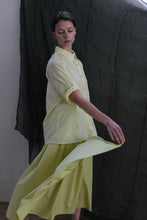 Load image into Gallery viewer, A person models the Work Shirt | Textured Lawn Citrine, paired with a matching flowing skirt, posing gracefully before a dark, sheer fabric backdrop.
